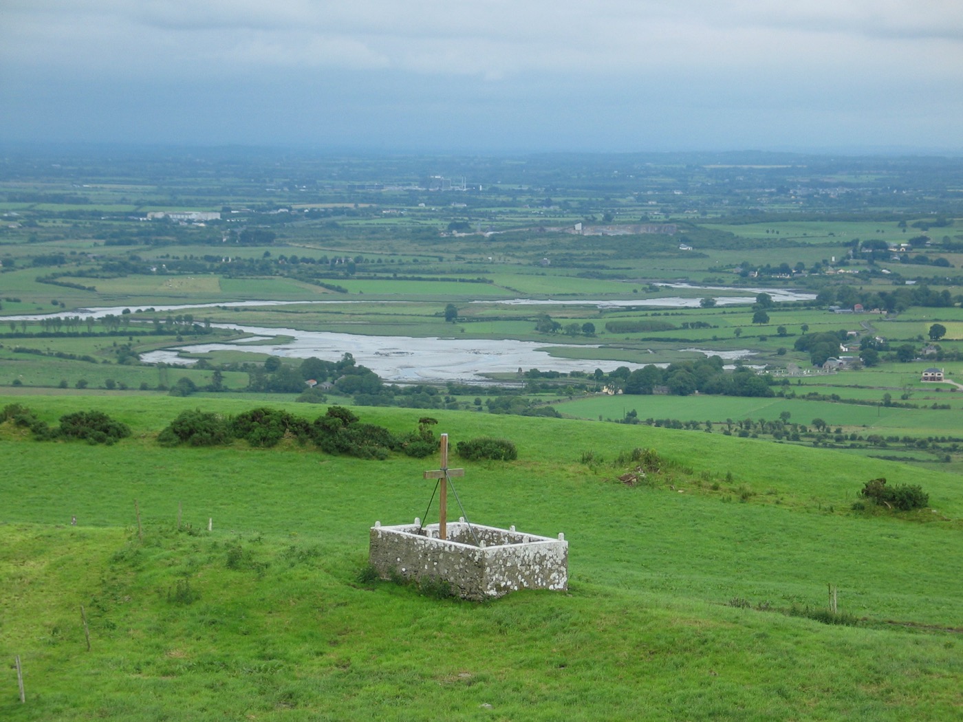 A white-walled hilltop enclosure with a wooden cross overlooking a green river valley.