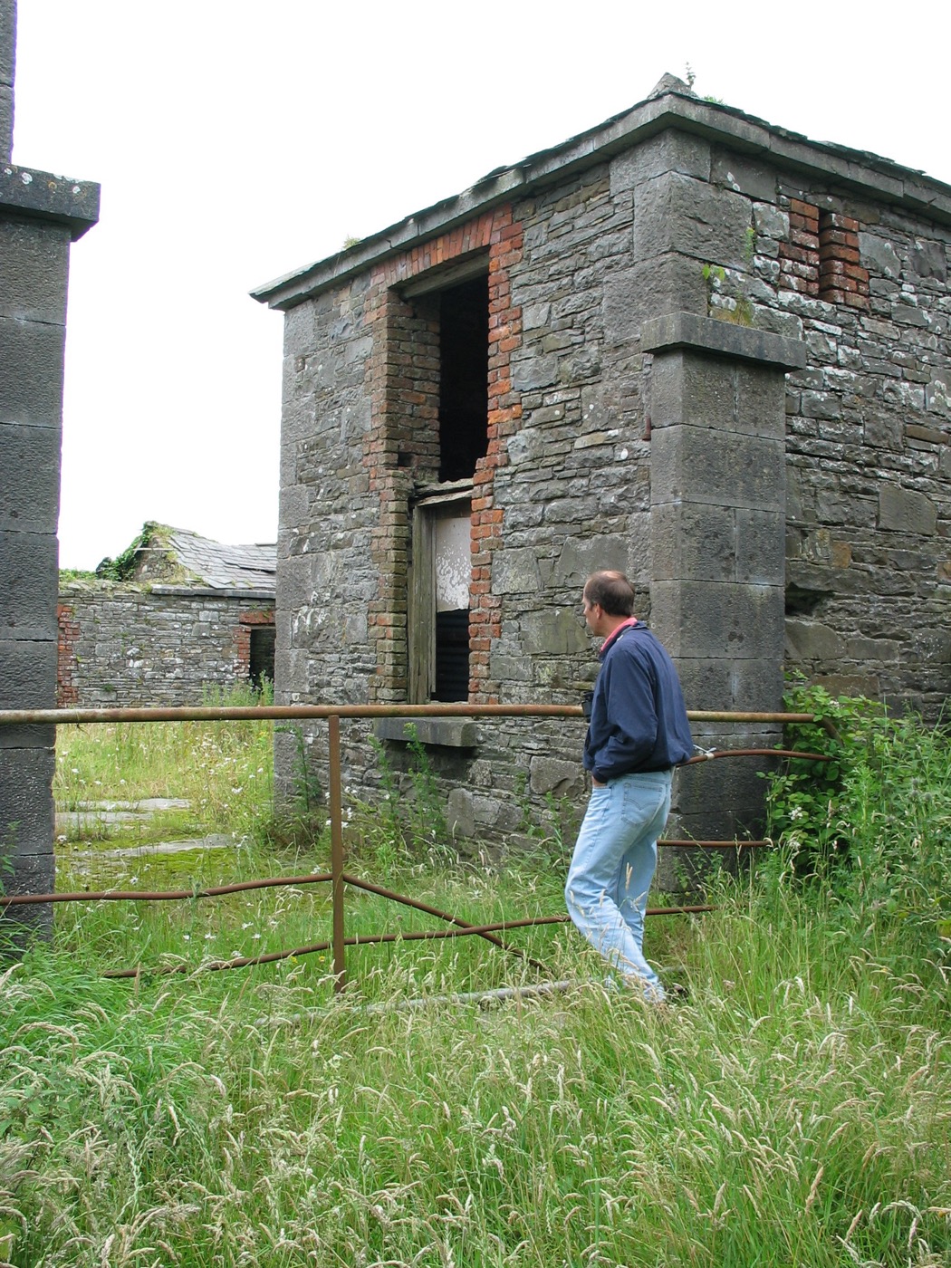 A man standing beside a ruined stone outbuilding with red-brick doorway dressings.